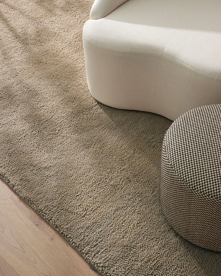 Neutral minimalist living room with cream curved furniture on a textured beige wool rug, layered with a patterned pouf, above pale timber flooring