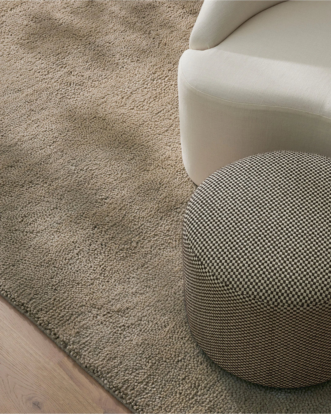 Neutral minimalist interior with cream upholstered chair and patterned ottoman on textured wool rug, showcasing layered beige tones and woven textures