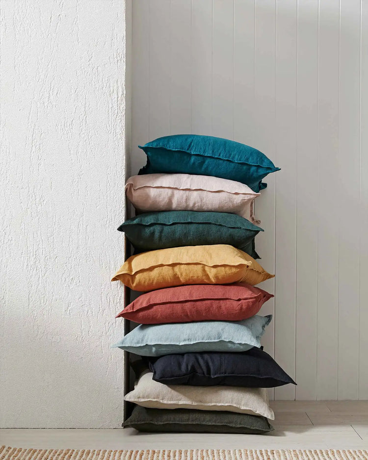 Stacked linen cushions in teal, cream, forest, gold, terracotta, blue and charcoal tones against textured white walls, showcasing neutral and earthy colour palette for modern home décor styling