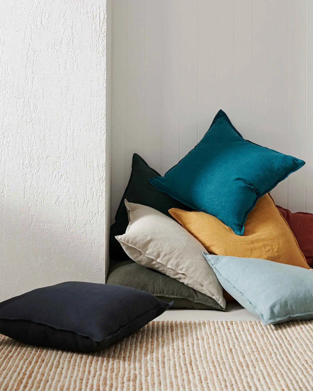 Layered linen cushions in teal, ochre, cream and navy create a contemporary colour study on neutral flooring, blending deep jewel tones with warm and cool neutrals against textured white walls.