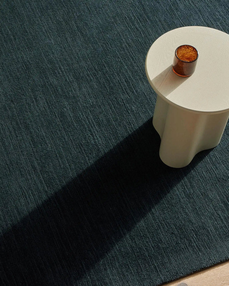 Teal wool rug with linear texture beneath a white side table and amber glass, minimalist interior styling with natural shadow play