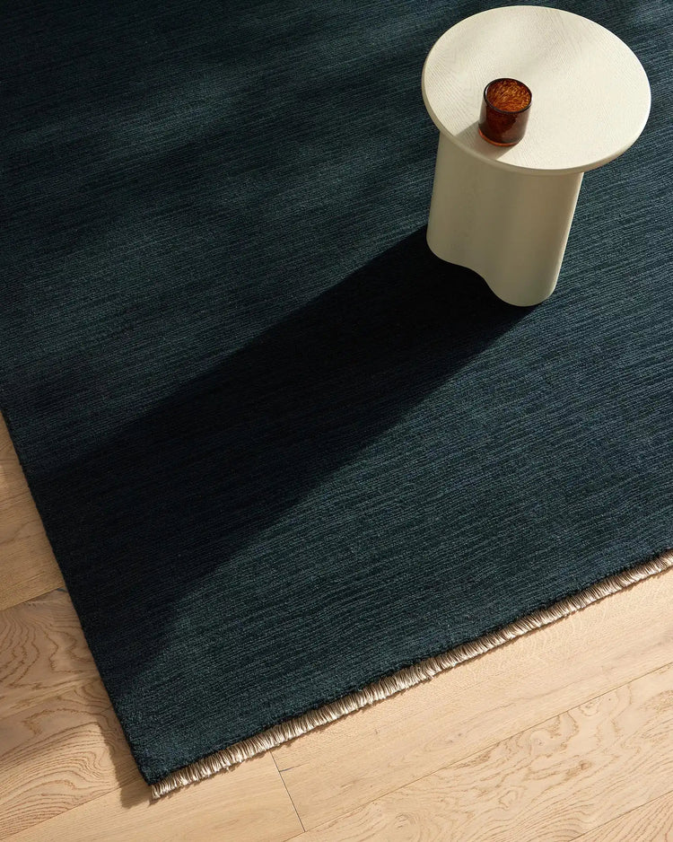 Deep teal wool rug with woven texture in minimalist room setting, styled with white pedestal table and amber glass accent on light wood flooring