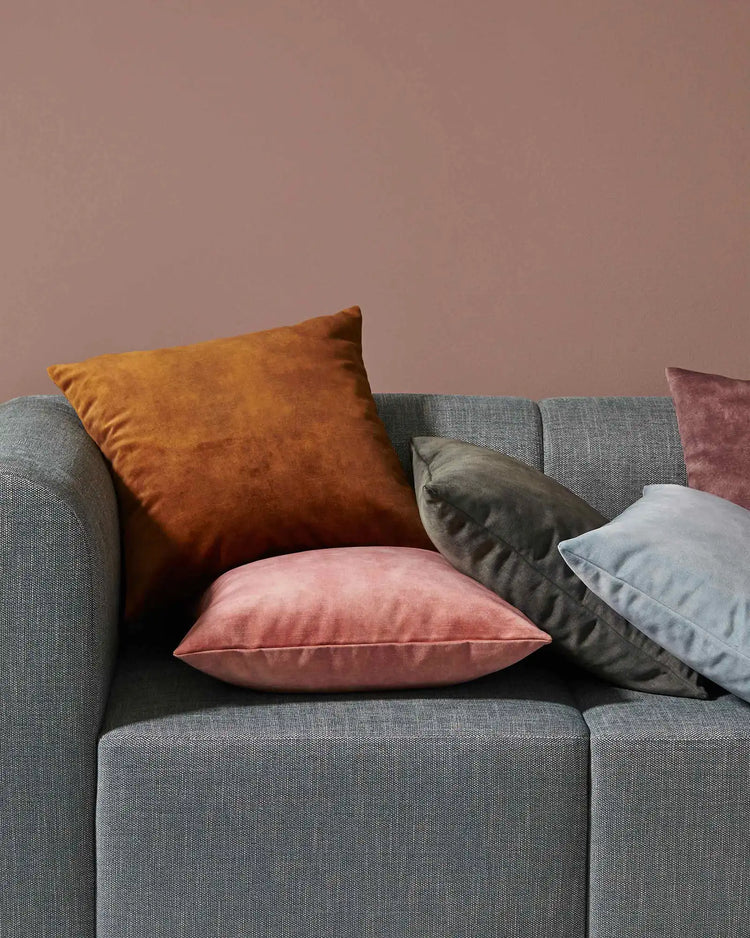 Warm earth tones and soft jewel hues styled on grey upholstered sofa with velvet Ava cushions in ochre, blush, charcoal and powder blue against mauve wall