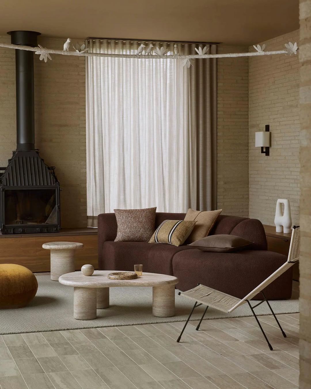Warm-toned living room styled with chocolate brown sofa, neutral linen curtains, and layered cushions in taupe, tan and terracotta. Textured wool rug anchors space with travertine and timber accents.
