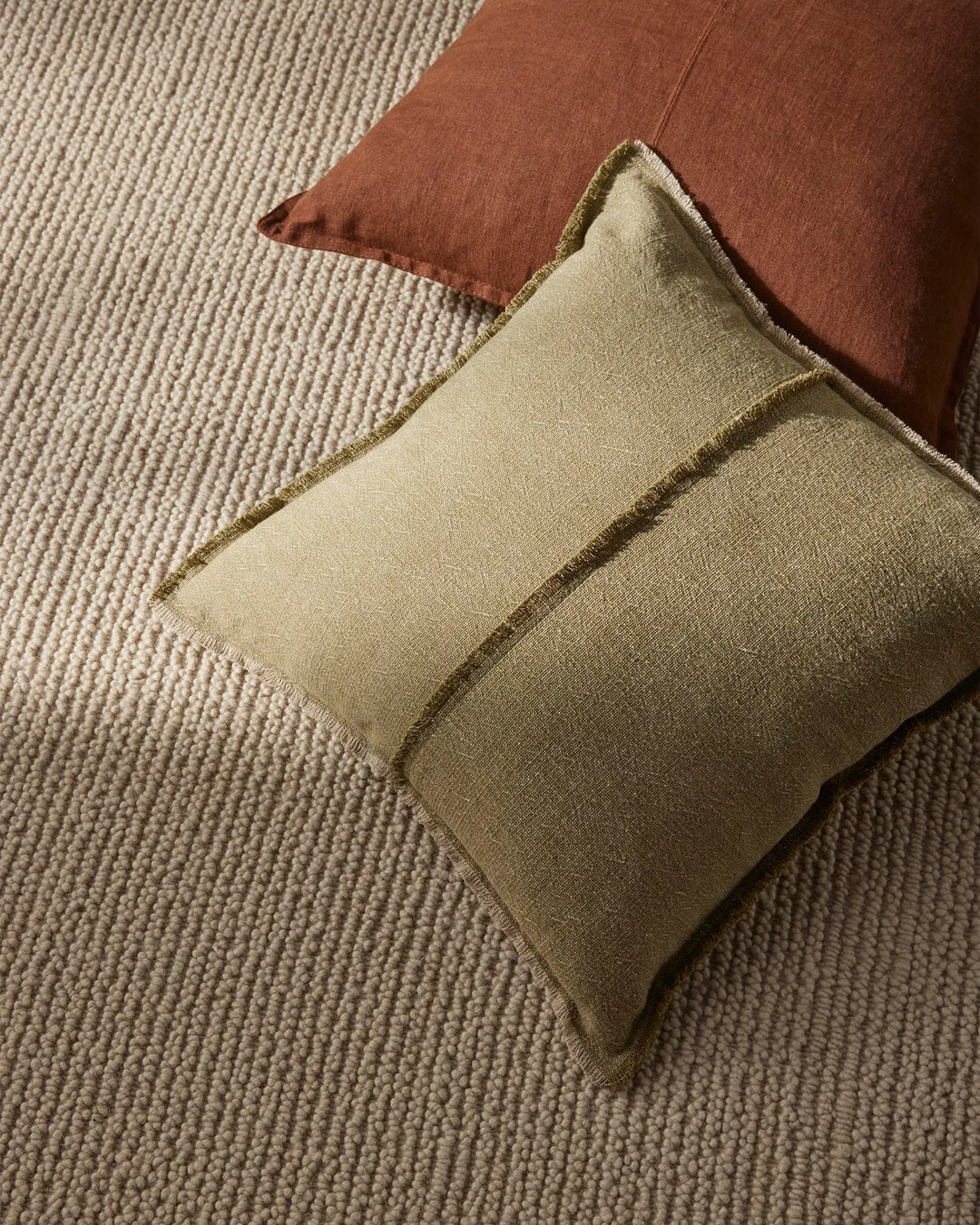 Warm earthy tones styled on neutral textured rug: sage linen cushion with whipstitched edge and rust-terracotta cushion, showcasing organic woven palette and tactile layering