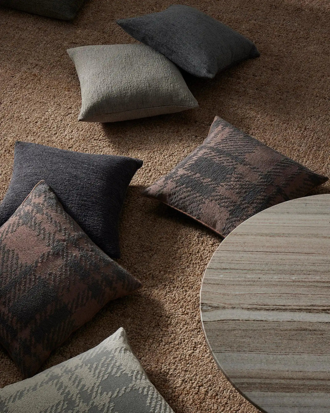 Neutral pillow collection on textured beige carpet featuring cream, charcoal, and taupe tones with geometric and solid weaves beside light wood table edge