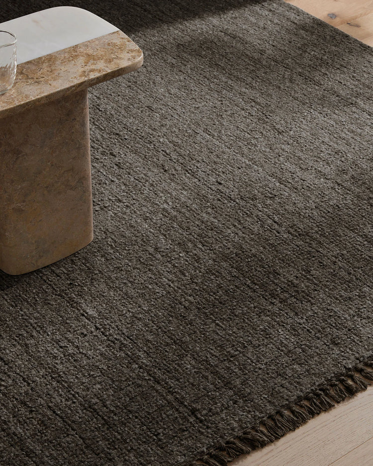 Charcoal wool-blend rug with fringed edge anchors minimalist room with travertine side table and light wood flooring