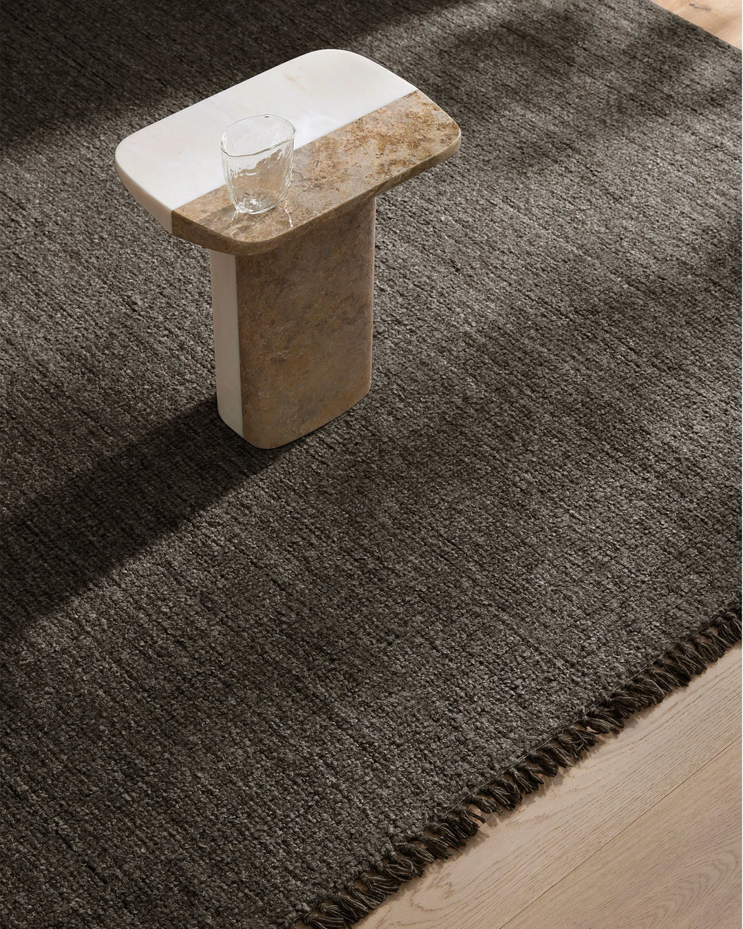 Charcoal wool-blend rug with fringe edge anchors minimalist space, styled with travertine side table and glass tumbler