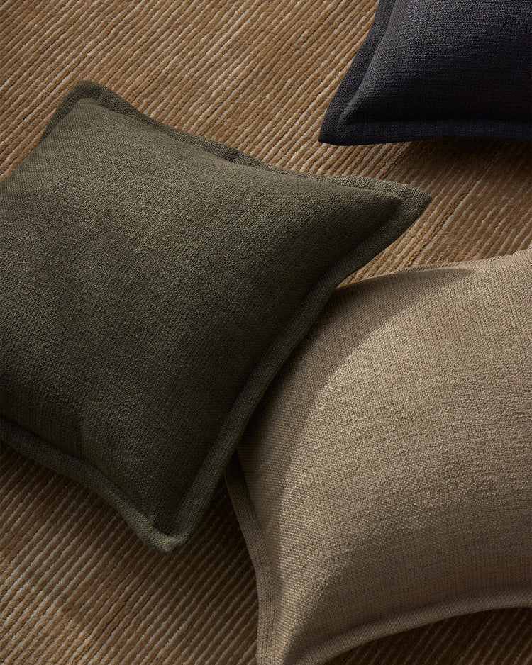 Earthy textured cushions in sage and taupe arranged on a warm wool rug, creating a layered neutral palette with soft tonal contrast