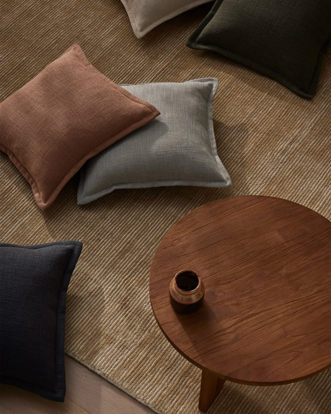 Warm, earthy interior styling with Figlio cushions in terra, kale, and mineral tones arranged on a textured wool rug beside a wooden round side table