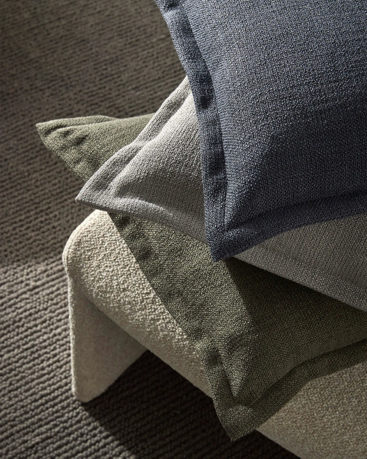 Neutral toned cushions in charcoal, sage and cream stacked on light wood chair against textured carpet, showcasing layered earth palette and boucle fabric texture