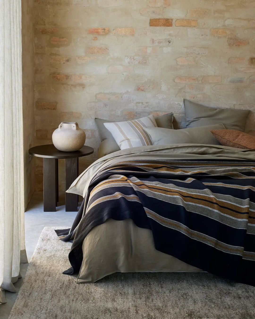 Minimalist bedroom with exposed brick wall, neutral bedding layered with Franco throw in navy and ochre stripes, linen pillows in soft taupe, wooden side table with ceramic vessel
