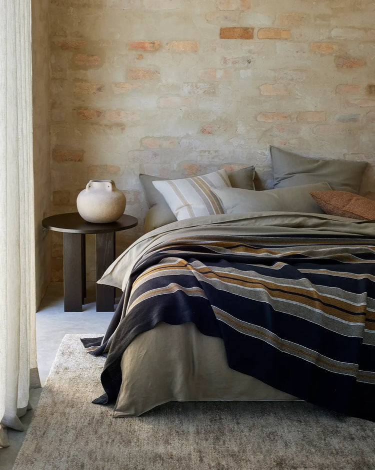 Minimalist bedroom with exposed brick wall, neutral bedding layered with Franco throw in navy and ochre stripes, linen pillows in soft taupe, wooden side table with ceramic vessel
