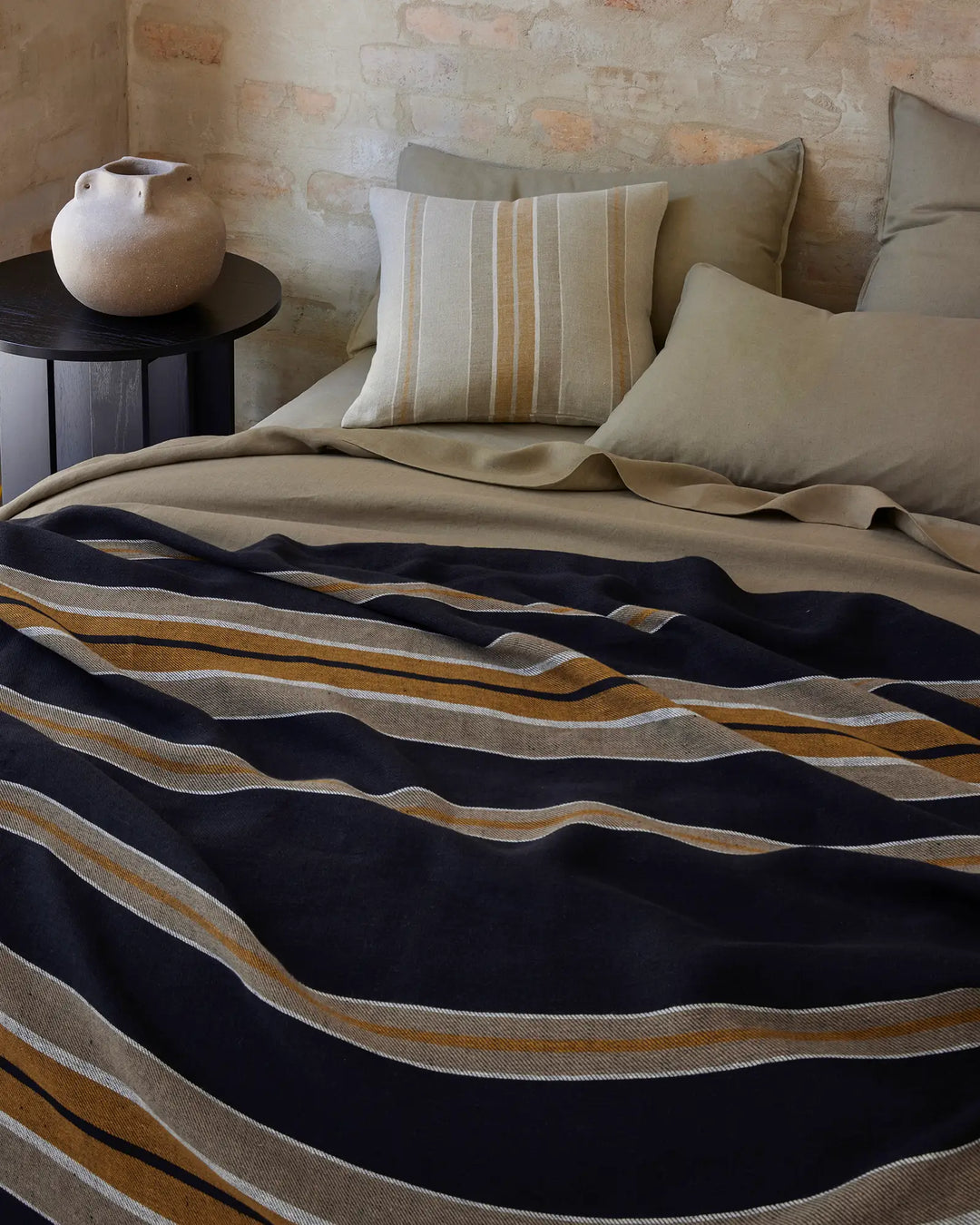Neutral bedroom with textured brick wall, layered beige bedding and striped cushion, Franco throw in navy, ochre and cream stripe drape across bed
