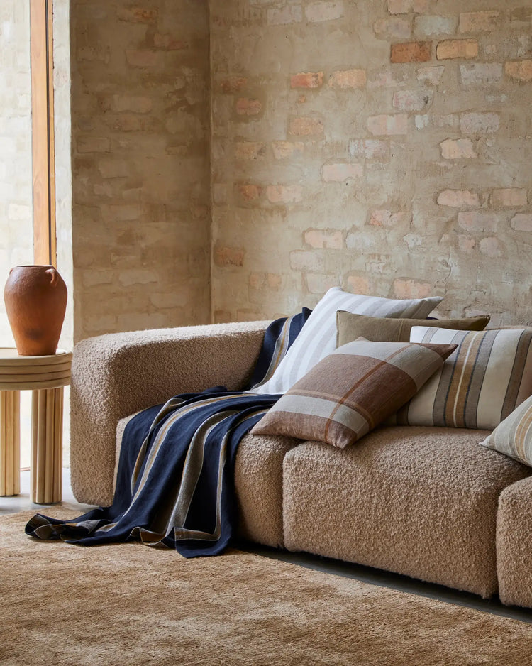 Neutral linen throw draped over a textured beige sofa with striped pillows, against exposed brick wall in warm earthy tones
