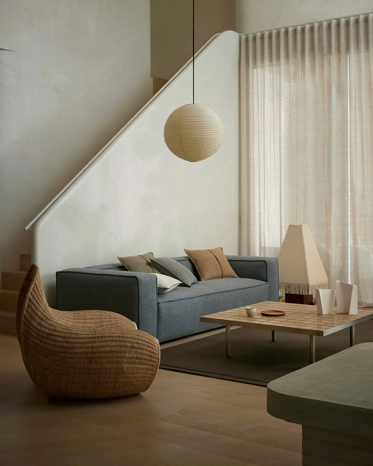 Minimalist living room with grey sofa layered with Adela and Alba cushions in earthy tones, woven chair, wooden coffee table, and Cassio wool rug in soft neutral palette with linen curtains and pendant lighting