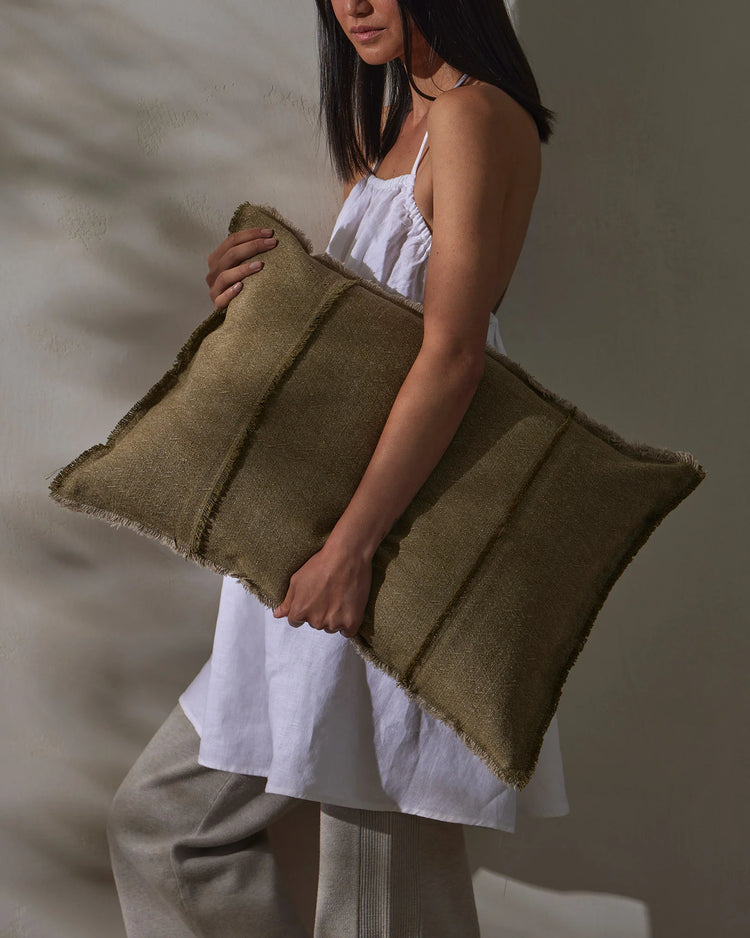Sage green linen cushion with raw frayed edges, styled against neutral minimalist backdrop with white cotton layers and soft natural light casting shadows