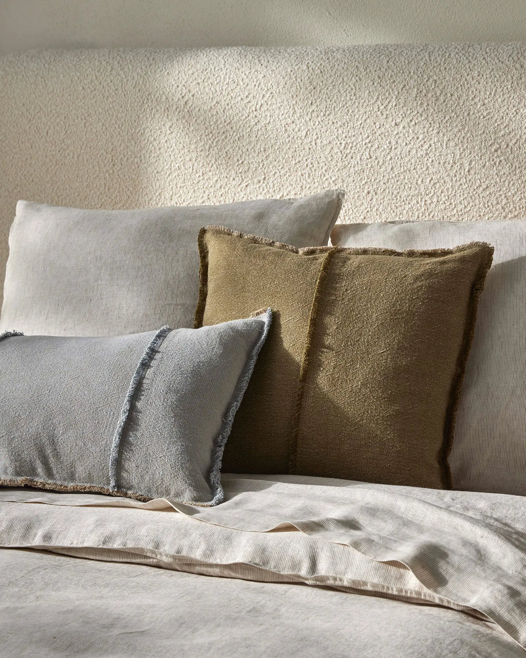 Neutral bedroom styling with layered cushions in sage, olive, and warm sand tones, textured linen bedding, and soft natural light on textured walls