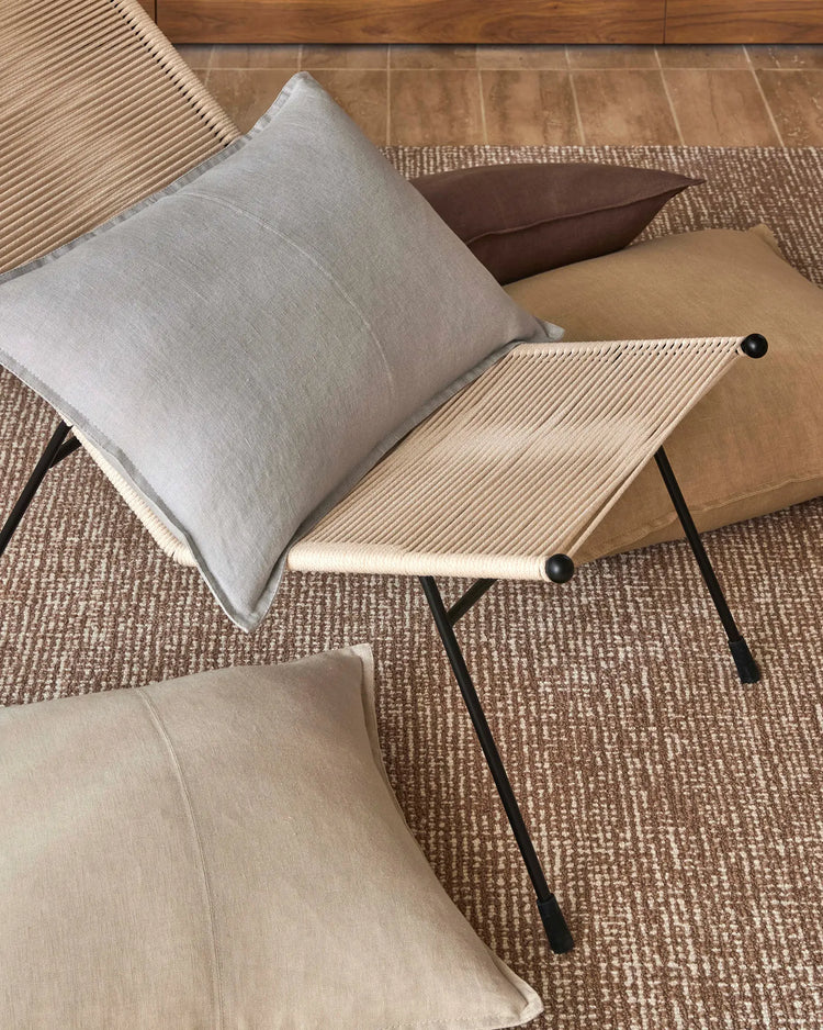 Neutral linen cushions arranged on woven jute rug with mid-century modern furniture; earthy palette of beige, cream and soft grey with black metal accents