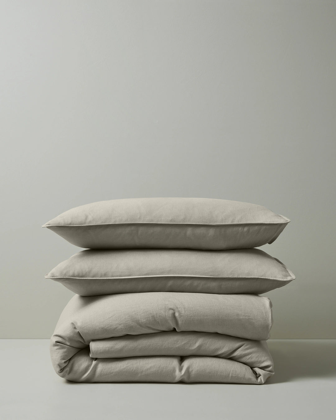Stacked natural linen bedding in cream and soft sage tones featuring Ravello quilt cover and pillowcase set, showcasing woven texture and relaxed, modern minimalist aesthetic against neutral background