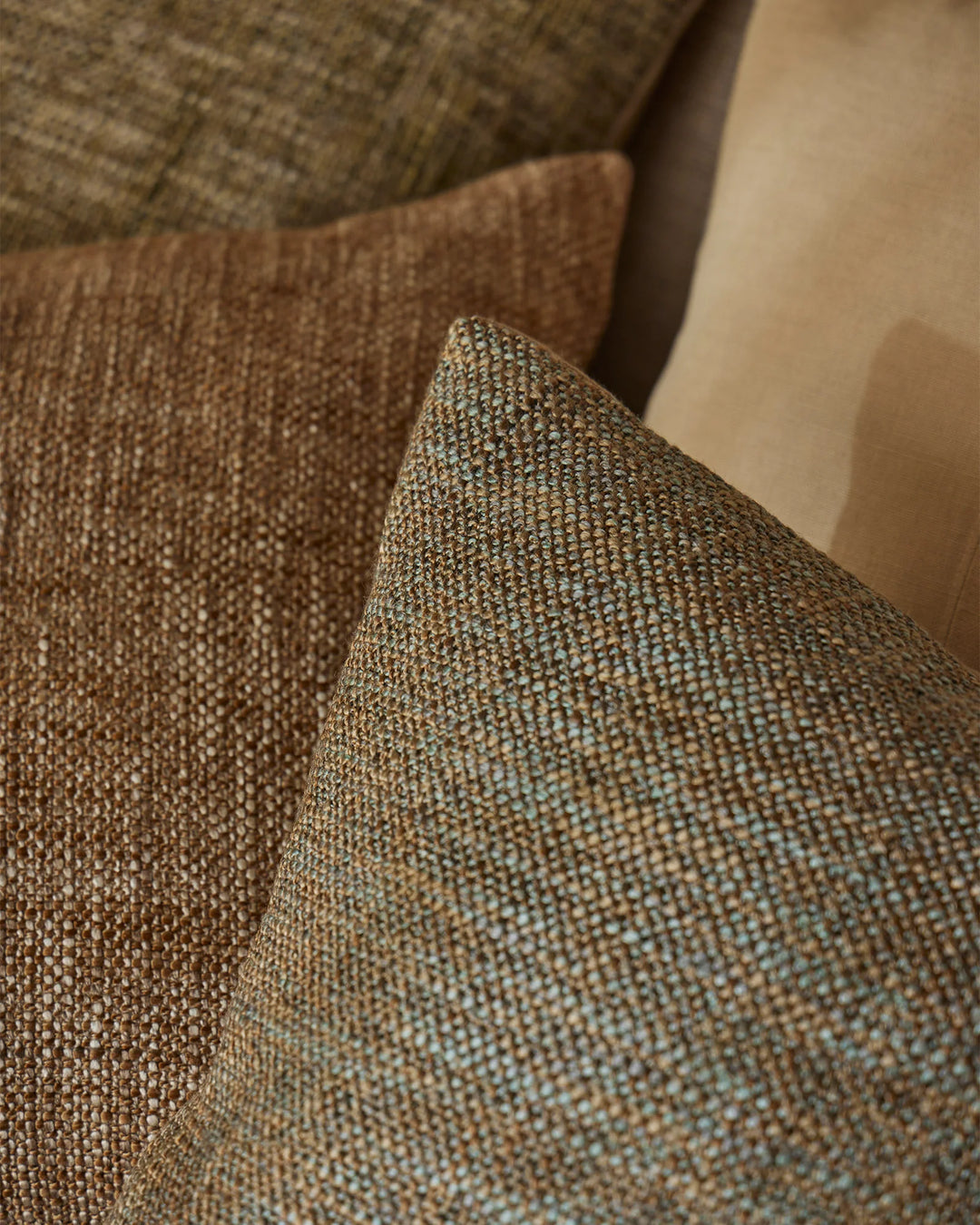 Earthy textured cushions in warm toffee and moss tones layered on neutral upholstery, highlighting woven fabric detail and warm minimalist home styling