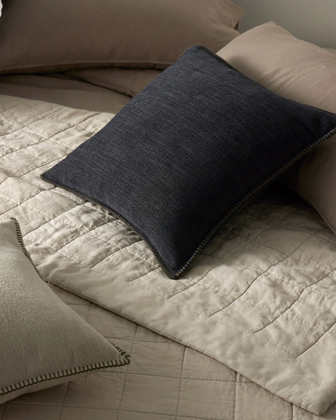 Neutral bed styling with cream quilted Ravello coverlet and charcoal linen Alba cushion, showcasing minimalist monochromatic palette and layered textures