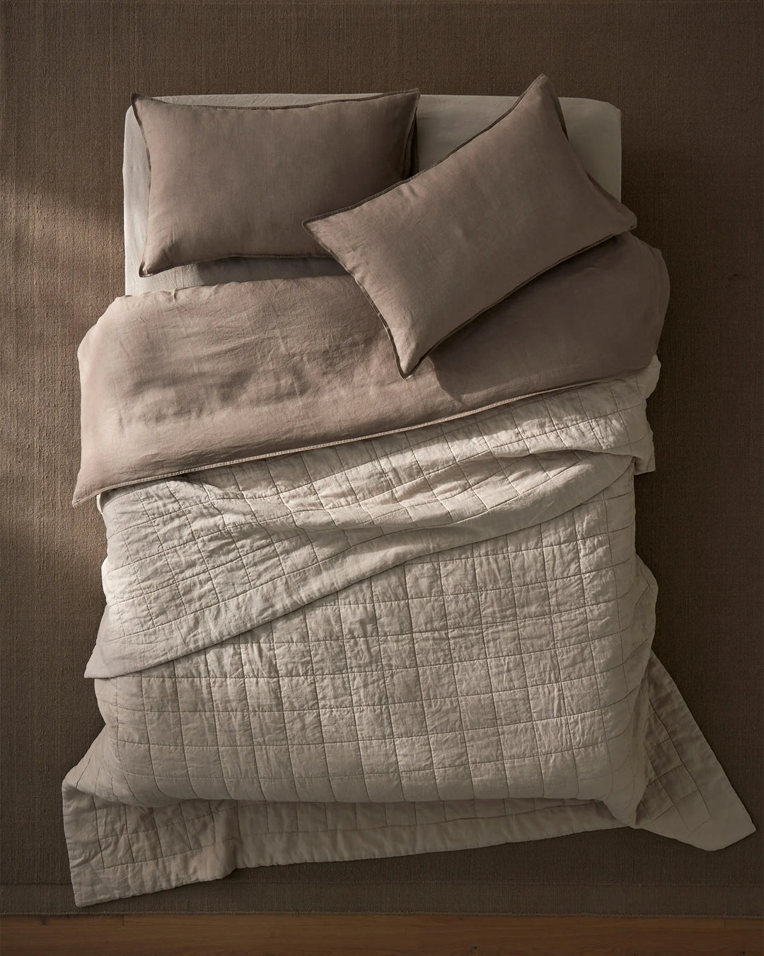 Overhead view of a neutral linen bed with Ravello Coverlet and Pillowcase Set in bone and pebble tones, layered with quilted textures and soft natural light casting shadows on warm taupe walls