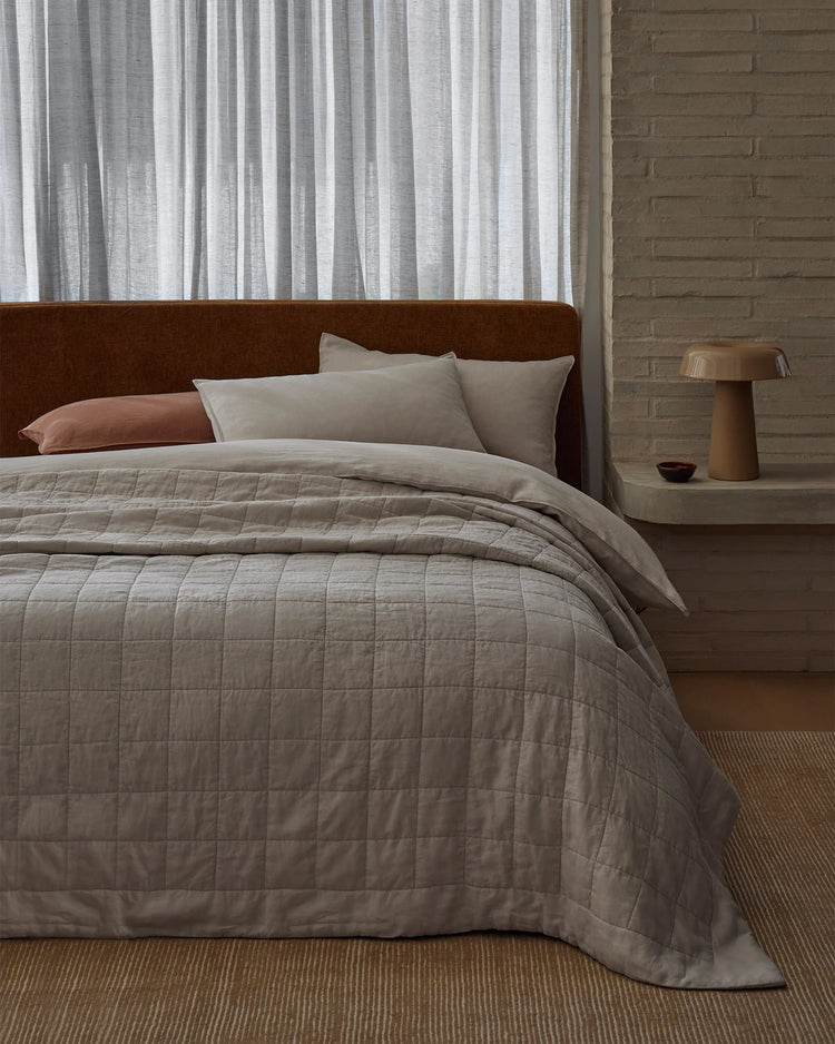 Minimalist bedroom with bone linen coverlet layered over neutral bedding, terracotta accent pillow, brown suede headboard, and sheer striped curtains in contemporary earthy palette