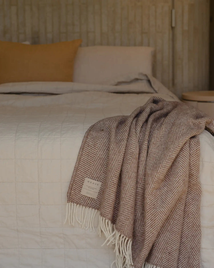Neutral bedroom styled with herringbone wool throw draped over quilted linen coverlet, warm terracotta cushion, and natural linen curtains creating a warm minimalist aesthetic