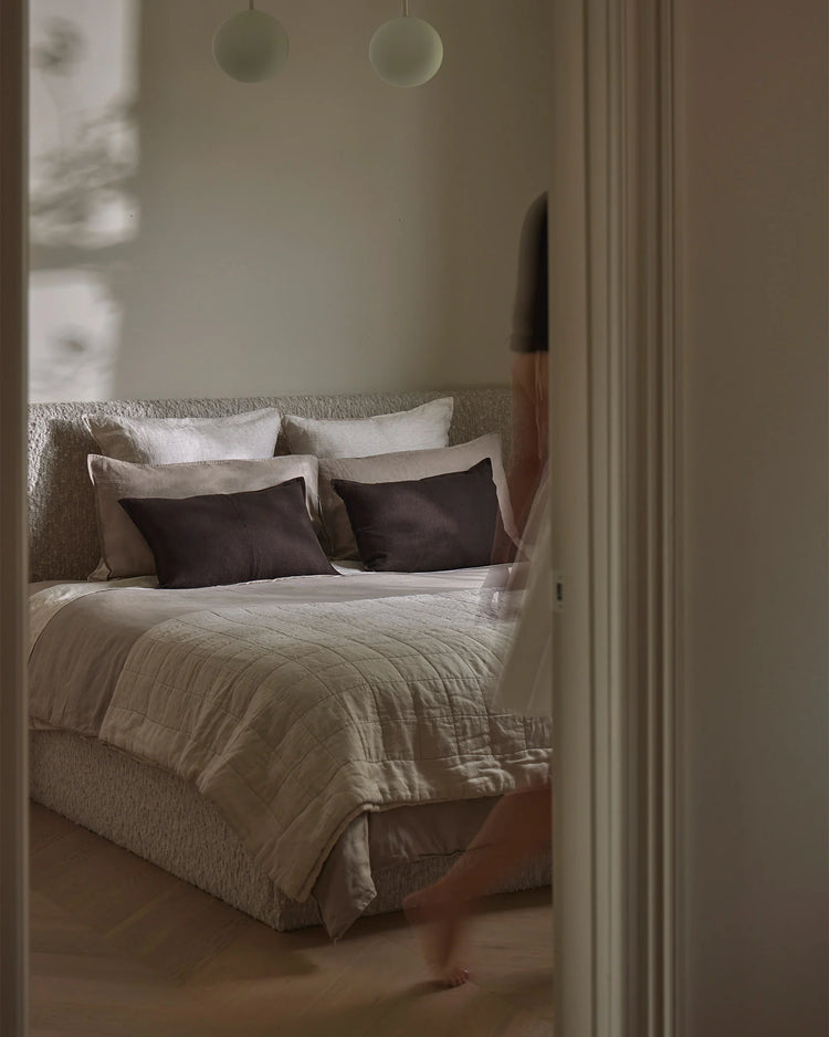 Minimalist bedroom styled with neutral bedding in cream and charcoal, featuring quilted linen coverlet layered with soft pillows against natural linen headboard, muted earthy palette with pendant lighting