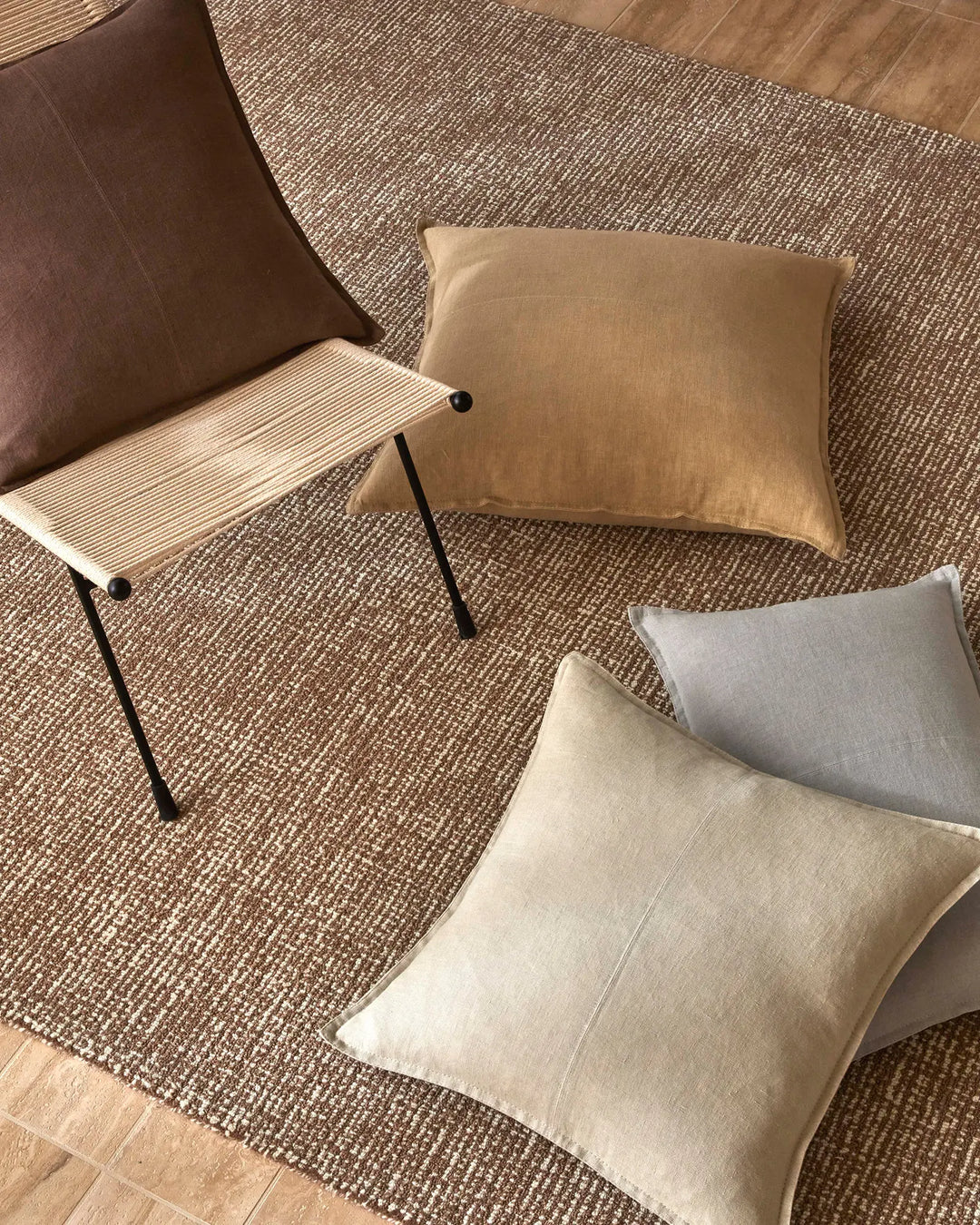 Neutral living room styled with Como linen cushions in sandstone, oatmeal and cocoa tones layered on textured jute rug, beside mid-century chair and wooden flooring
