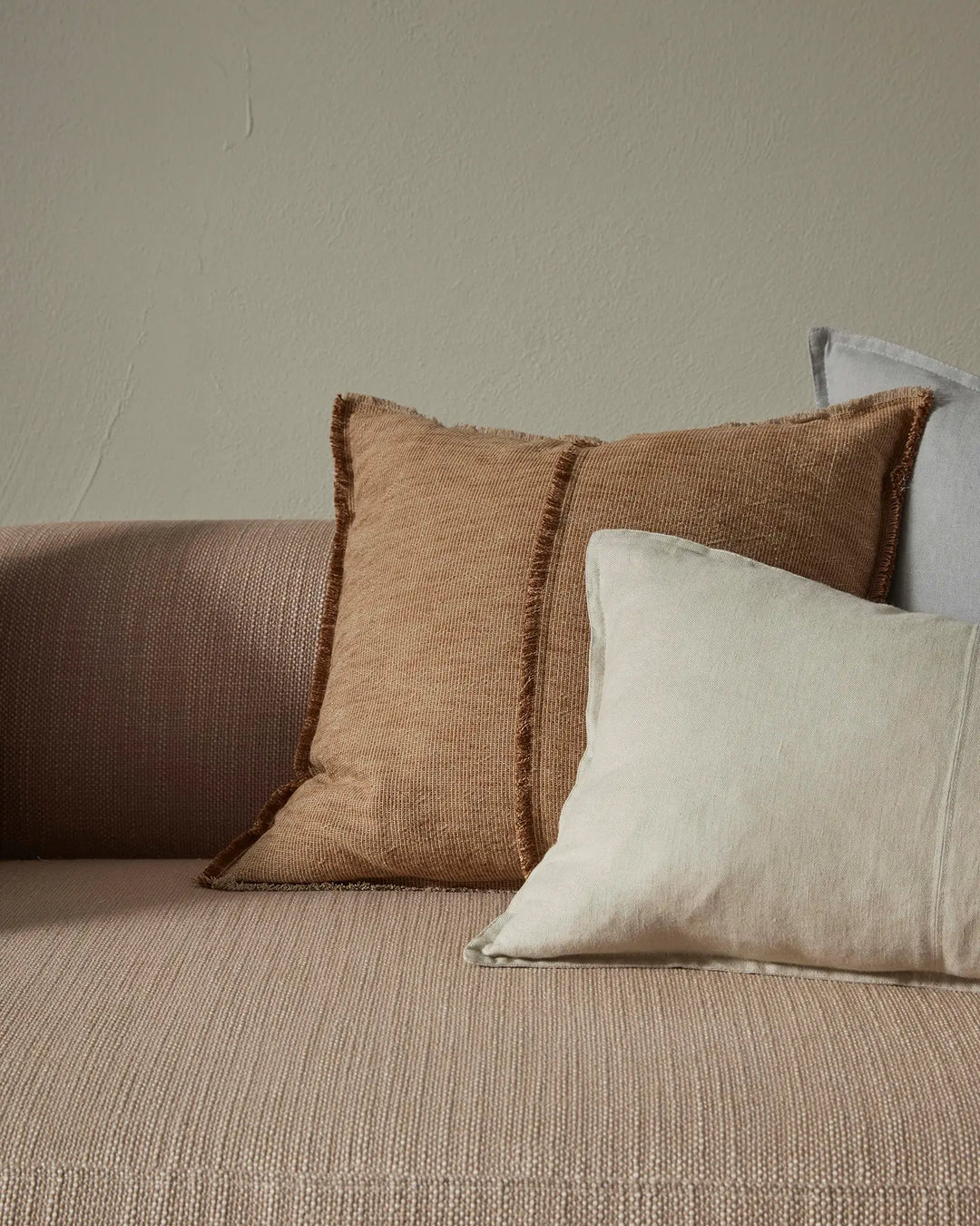 Neutral linen cushions in oatmeal and desert tones styled on a taupe sofa, showcasing textured weaves and frayed edge details against a minimal cream wall