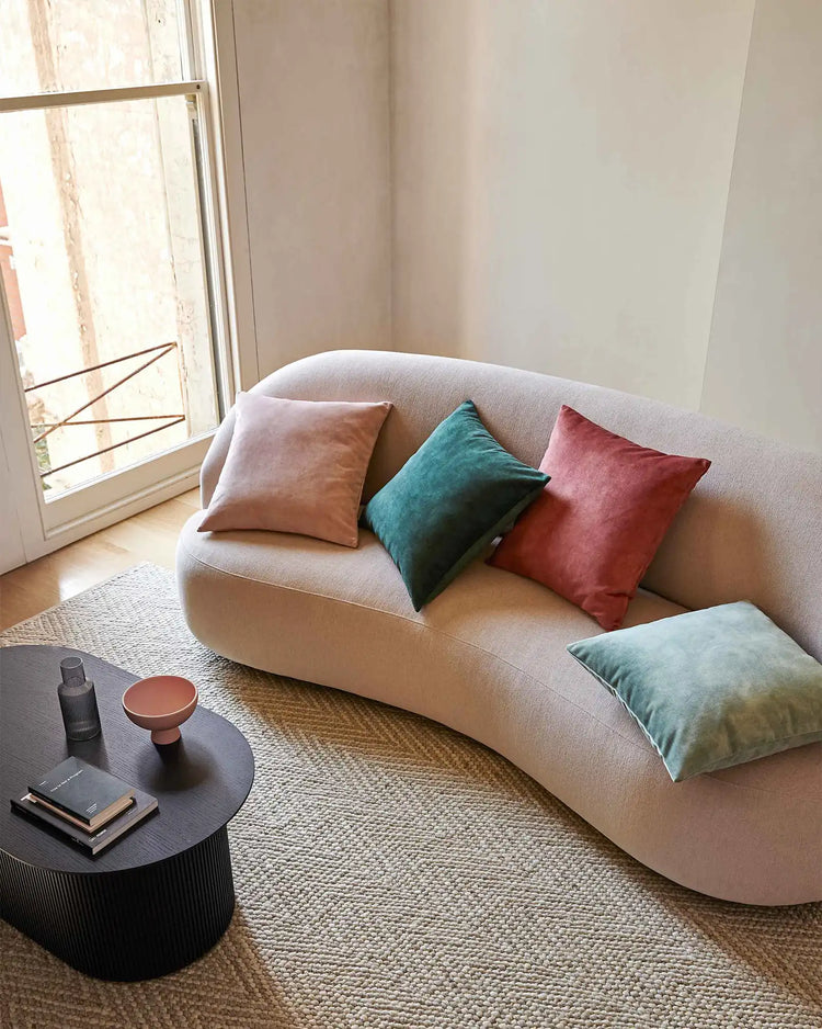 Curved cream sofa styled with jewel-tone and blush velvet cushions in a minimalist living room with natural light, neutral textures and warm wooden flooring