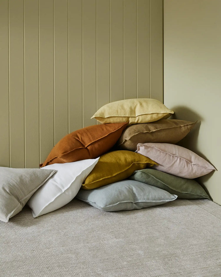 Stacked linen cushions in warm terracotta, mustard, blush and sage tones against a fluted olive green wall, creating an earthy, layered aesthetic with natural texture and neutral palette