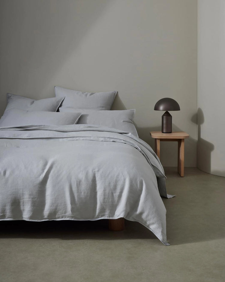 Minimalist bedroom with Ravello linen bedding in soft silver, crisp pillows, and natural wood nightstand with bronze mushroom lamp against neutral walls
