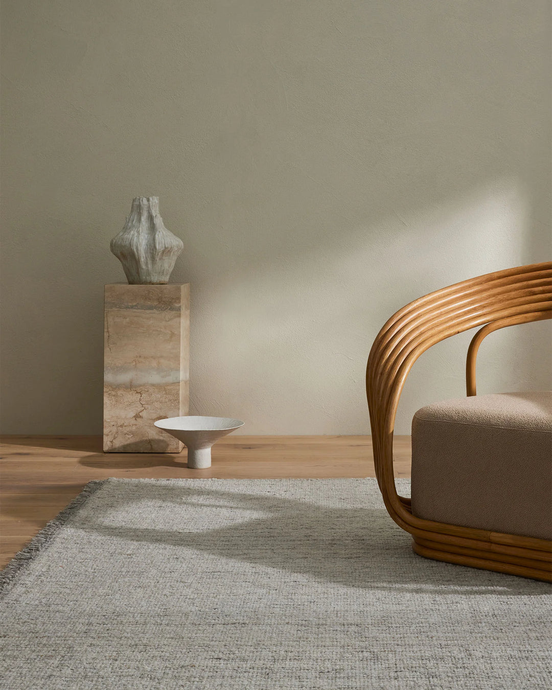 Warm minimalist room with neutral Puglia rug anchoring wooden chair and marble plinth, soft daylight casting geometric shadows on beige walls