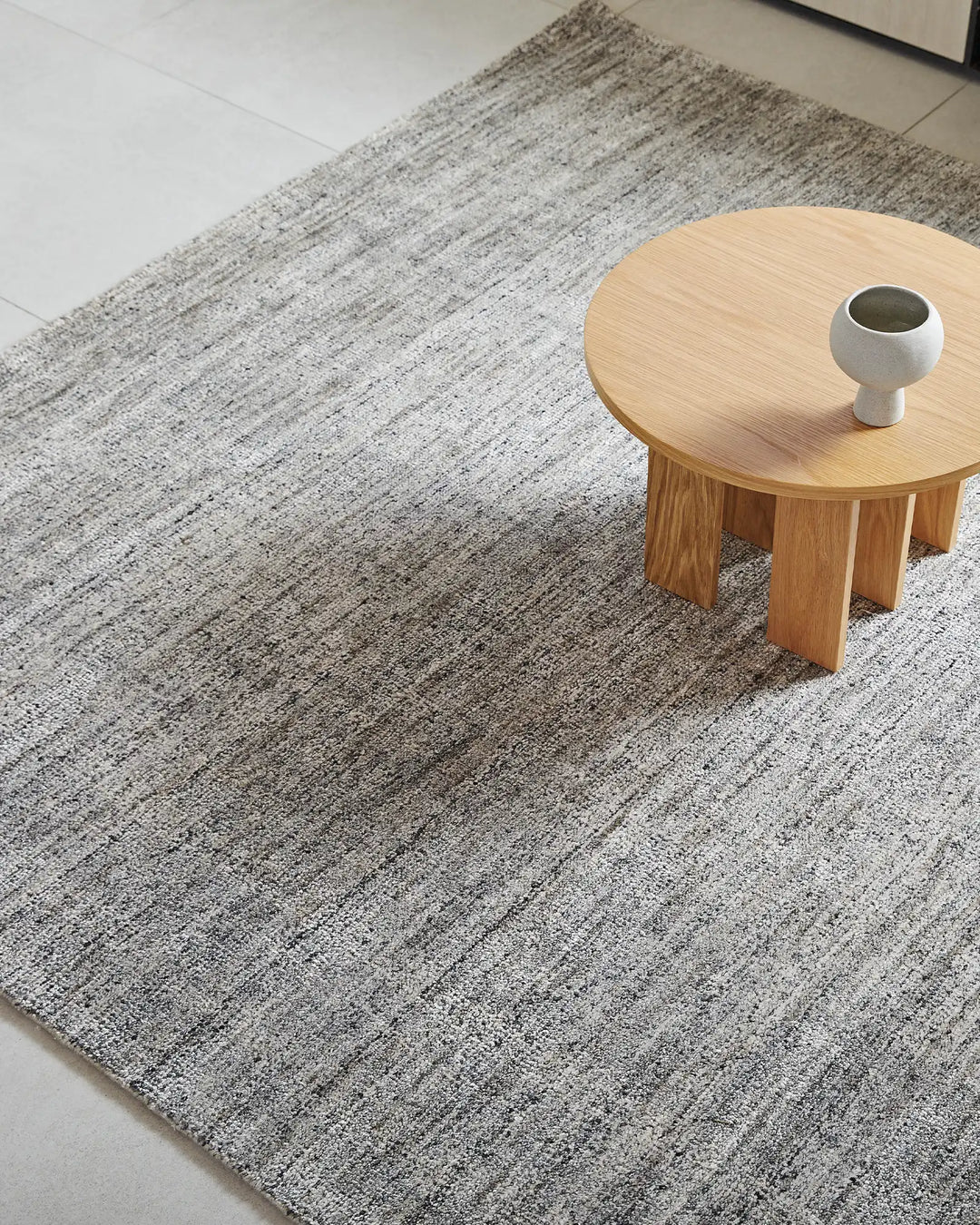 Granito rug in shale grey anchors a minimalist space with natural oak table and white ceramic vessel, showcasing textured wool-blend weave and linear pattern detail