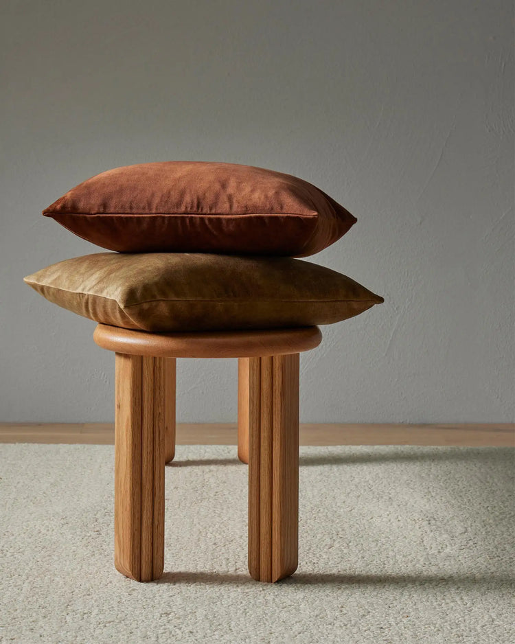 Stacked earth-tone cushions in rust and golden-brown on a wooden stool, styled against a neutral grey wall and natural fibre rug for warm, minimalist home décor