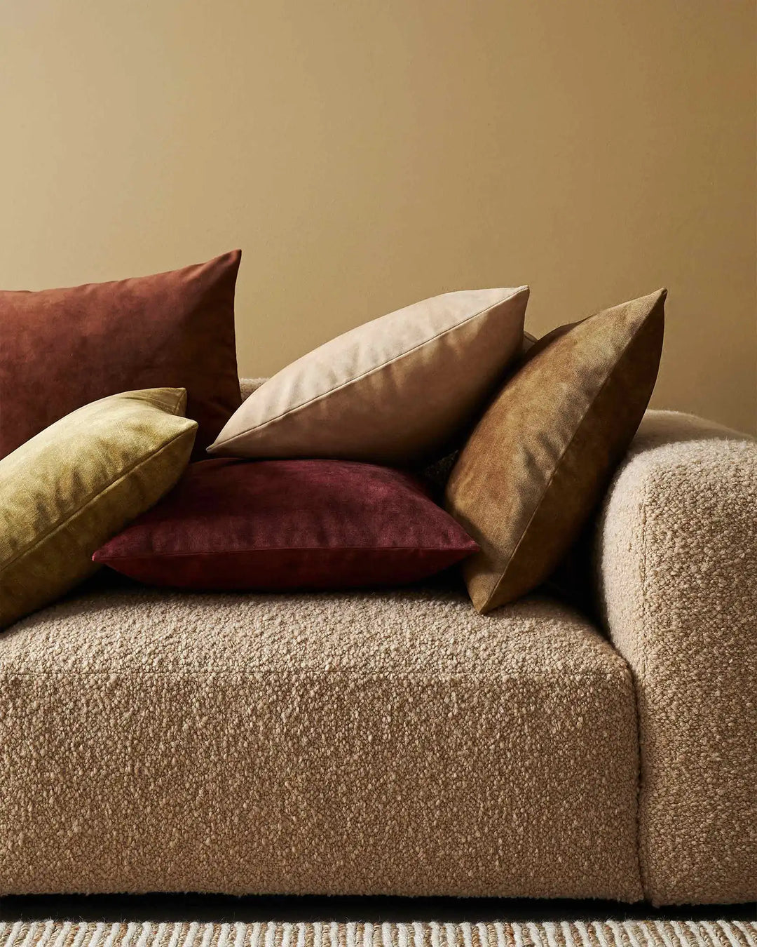 Warm earth-tone cushions in burgundy, gold, tan and cream arranged on a neutral textured sofa with beige wall and woven area rug backdrop