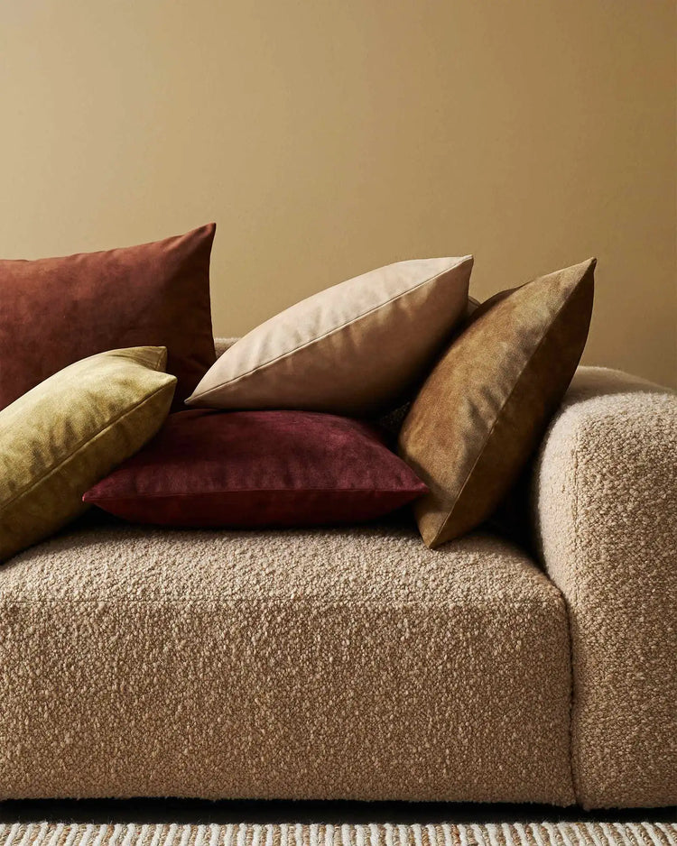Warm earth-tone cushions in burgundy, gold, tan and cream arranged on a neutral textured sofa with beige wall and woven area rug backdrop