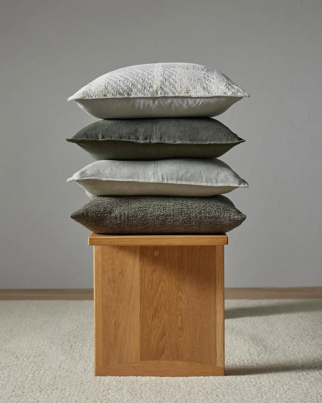 Stacked neutral cushions in cream, sage and charcoal tones showcase layered textures from linen and cotton blends, styled on a timber block base against soft grey backdrop
