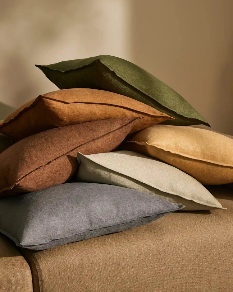 Stacked Fiore cushions in earthy tones—sage, olive, ochre, honey, toffee and denim—showcase linen viscose textures and natural warm lighting in neutral aesthetic interior styling.