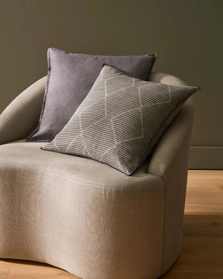 Neutral linen cushions in sage and charcoal on a curved cream armchair with timber flooring, creating a calm, textured living room aesthetic