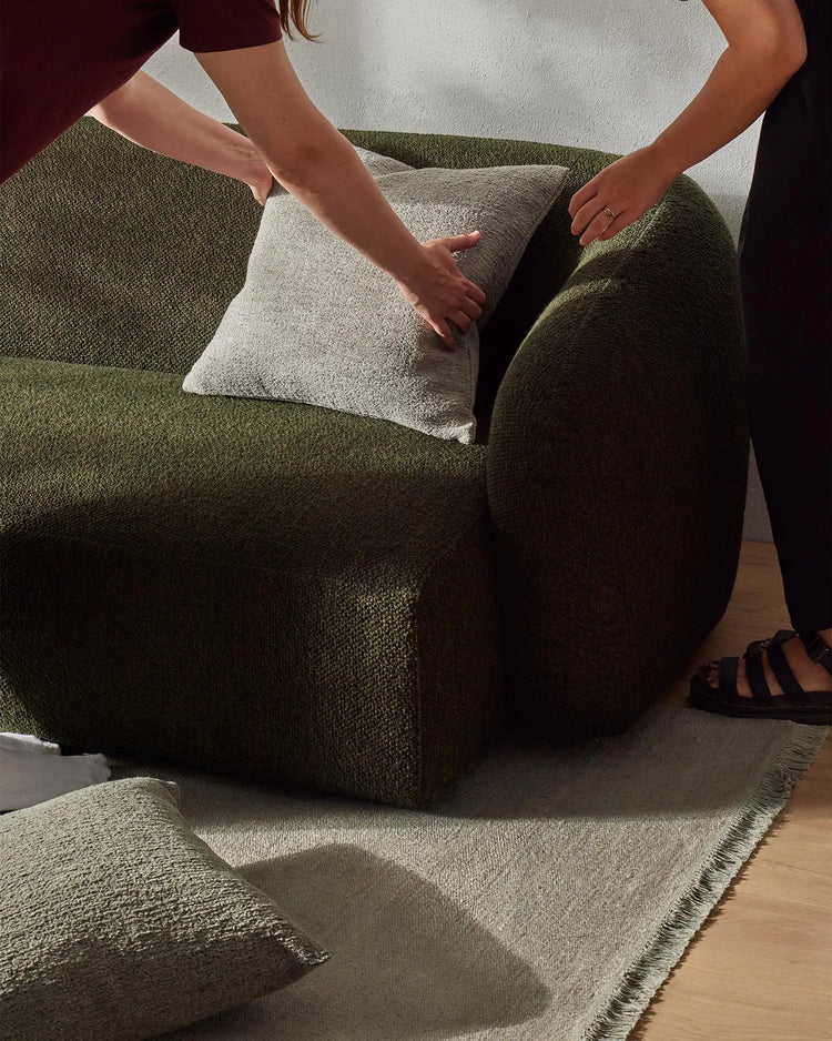 Earthy green and cream upholstered sofa with textured cushions and layered linen rug in neutral tones, hands touching woven fabric in casual living room setting