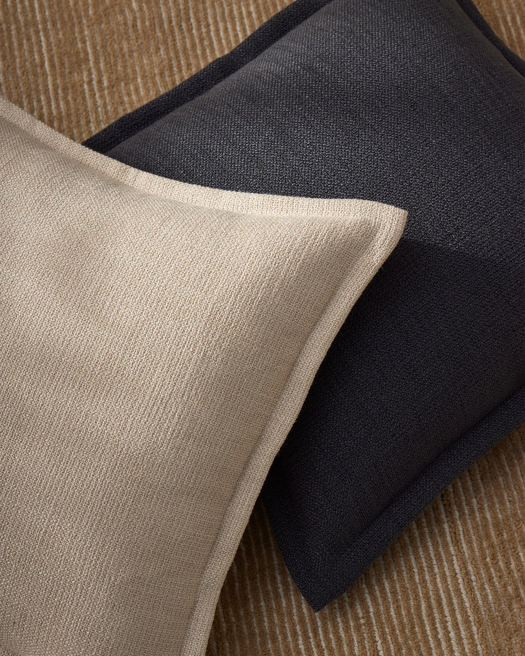 Neutral and charcoal cushions layered on textured cream rug, showcasing minimalist palette of warm beige, soft grey and deep charcoal with fine woven fabric textures