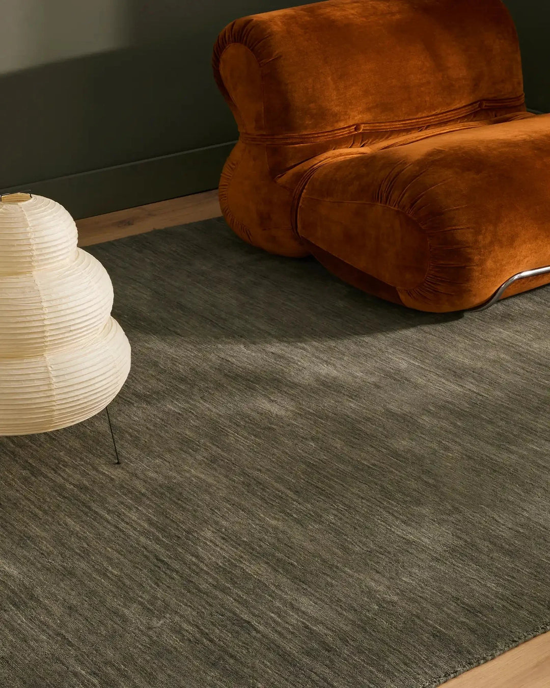 Charcoal wool-look rug anchors a mid-century seating vignette with cognac velvet armchair and cream pendant lamp against sage green wall