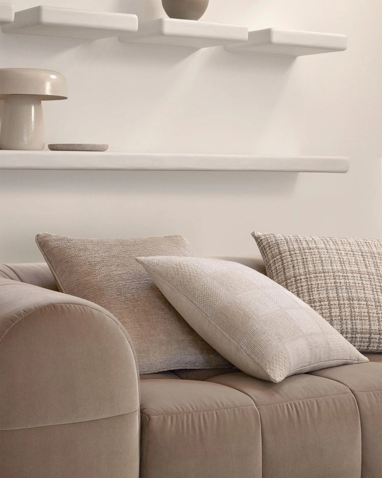 Neutral living room with cream curved sofa layered with Tempo, Bodrum and Tripoli cushions in oatmeal, hemp and fog tones, white shelving with ceramic vessels above