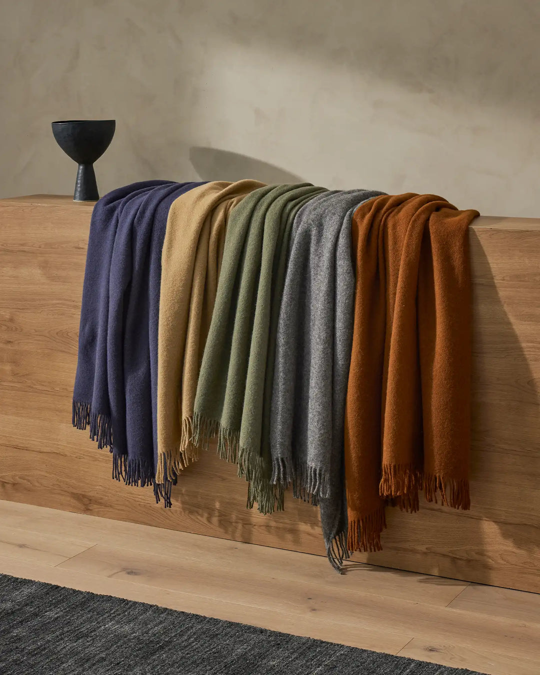 Nevis throws draped across wooden bench in navy, gold, sage, charcoal and rust tones, showcasing warm earth-toned wool palette against neutral plaster walls and natural wood flooring.