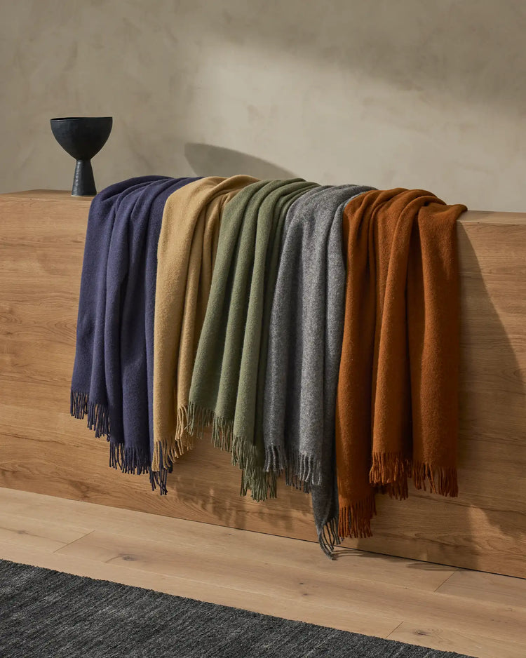 Nevis throws draped across wooden bench in navy, gold, sage, charcoal and rust tones, showcasing warm earth-toned wool palette against neutral plaster walls and natural wood flooring.