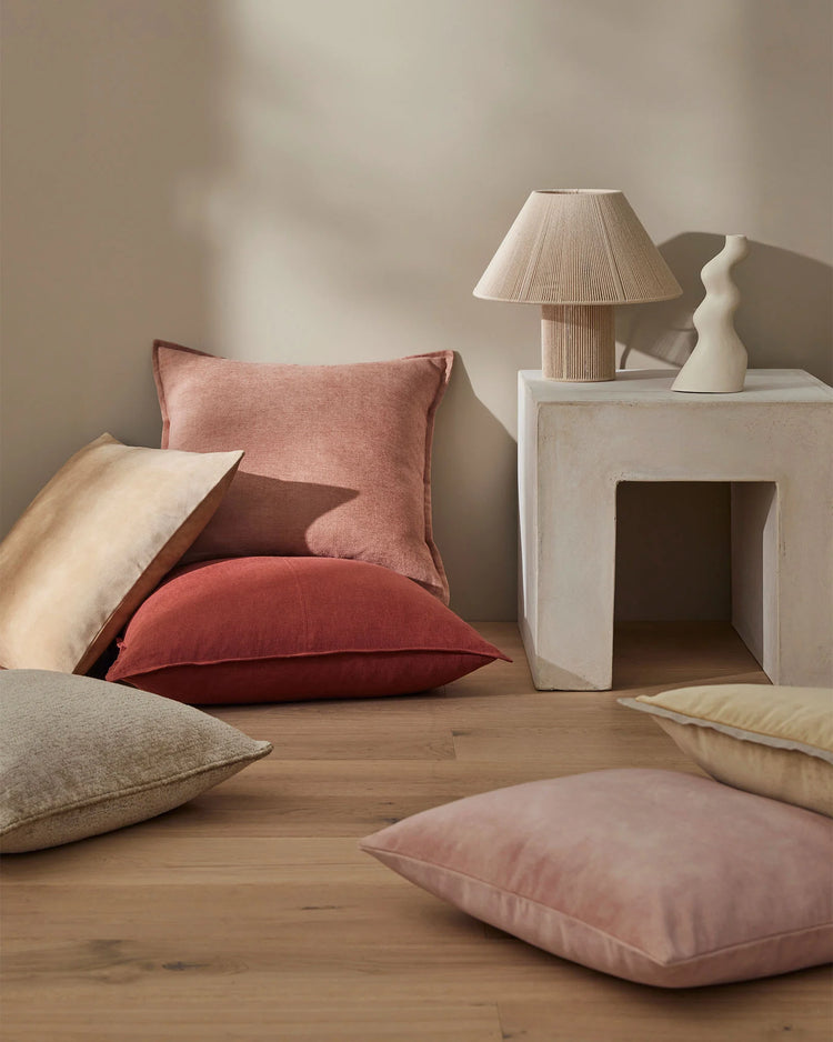 Warm earth-tone cushions in blush, burgundy, oatmeal and clay layered on wooden floor, styled with concrete plinth and textured lamp in minimalist Nordic interior